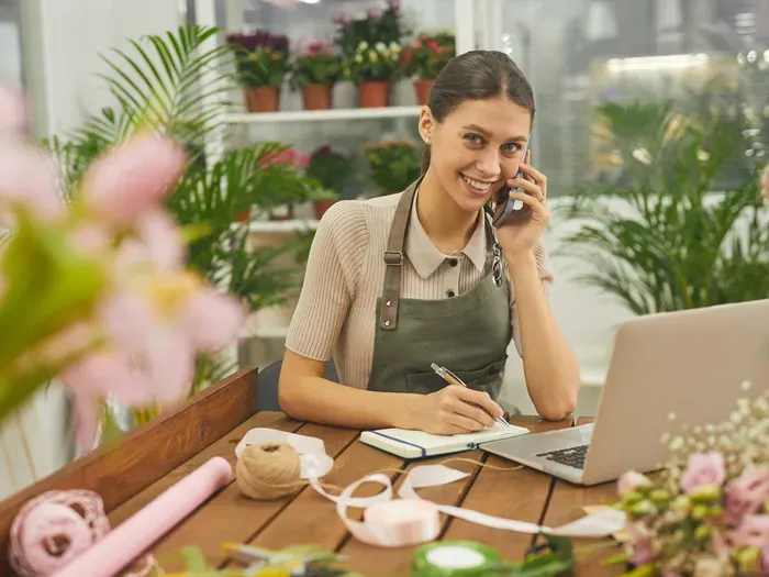 Smiling florist in an apron taking a phone order by a laptop, showing a local business listed on City Biz Zone